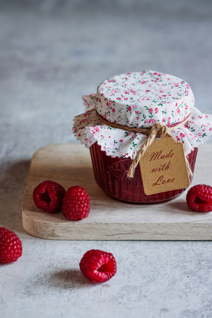 Samtige Himbeerkonfitüre ohne Kerne in einem auf einem Holzbrett mit Himbeeren im Vorder- und Hintergrund.