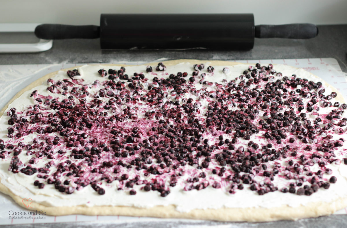 Ausgerollter Hefeteig für Hefeschnecken mit der verstrichenen Cheesecakefüllung und Blaubeeren.