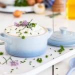 Kräuterquark mit Schnittlauch Petersilie und Schnittlauchblüten. Im Hintergrund Kartoffeln, Leinöl, Zwiebeln und Knoblauch sowie eine Pfeffermühle.