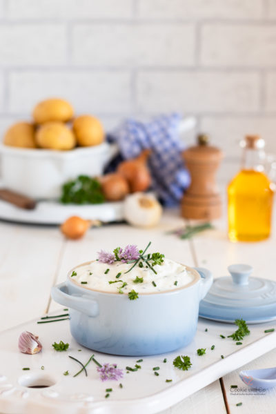 Kräuterquark mit Schnittlauch Petersilie und Schnittlauchblüten. Im Hintergrund Kartoffeln, Leinöl, Zwiebeln und Knoblauch sowie eine Pfeffermühle.