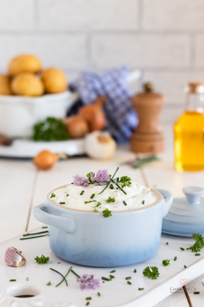 Kräuterquark mit Schnittlauch Petersilie und Schnittlauchblüten. Im Hintergrund Kartoffeln, Leinöl, Zwiebeln und Knoblauch sowie eine Pfeffermühle.