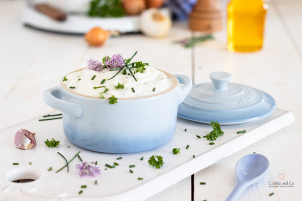 Quark mit Kraütern, wie Schnittlauch, Petersilie und Schnittlauchblüten. Im Hintergrund, Leinöl, Zwiebeln und Knoblauch sowie eine Pfeffermühle.