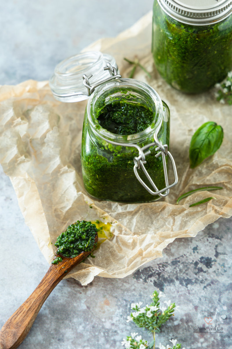 Mediterrane Kräuter-Gewürz-Paste auf einem Holzlöffel. Im Hintergrund frische Kräuter und ein Glas gefüllt mit der Paste.