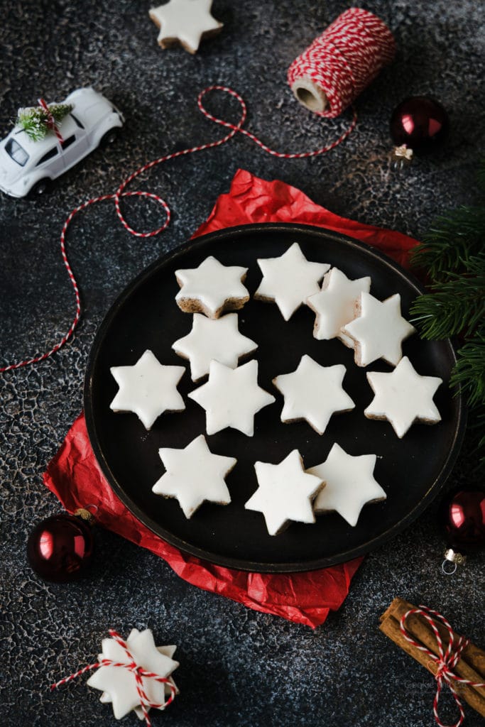 Zimtsterne auf einem Teller mit rotem Seidenpapier darunter und mit roten Weihnachtskugeln und Zimtstangen daneben.