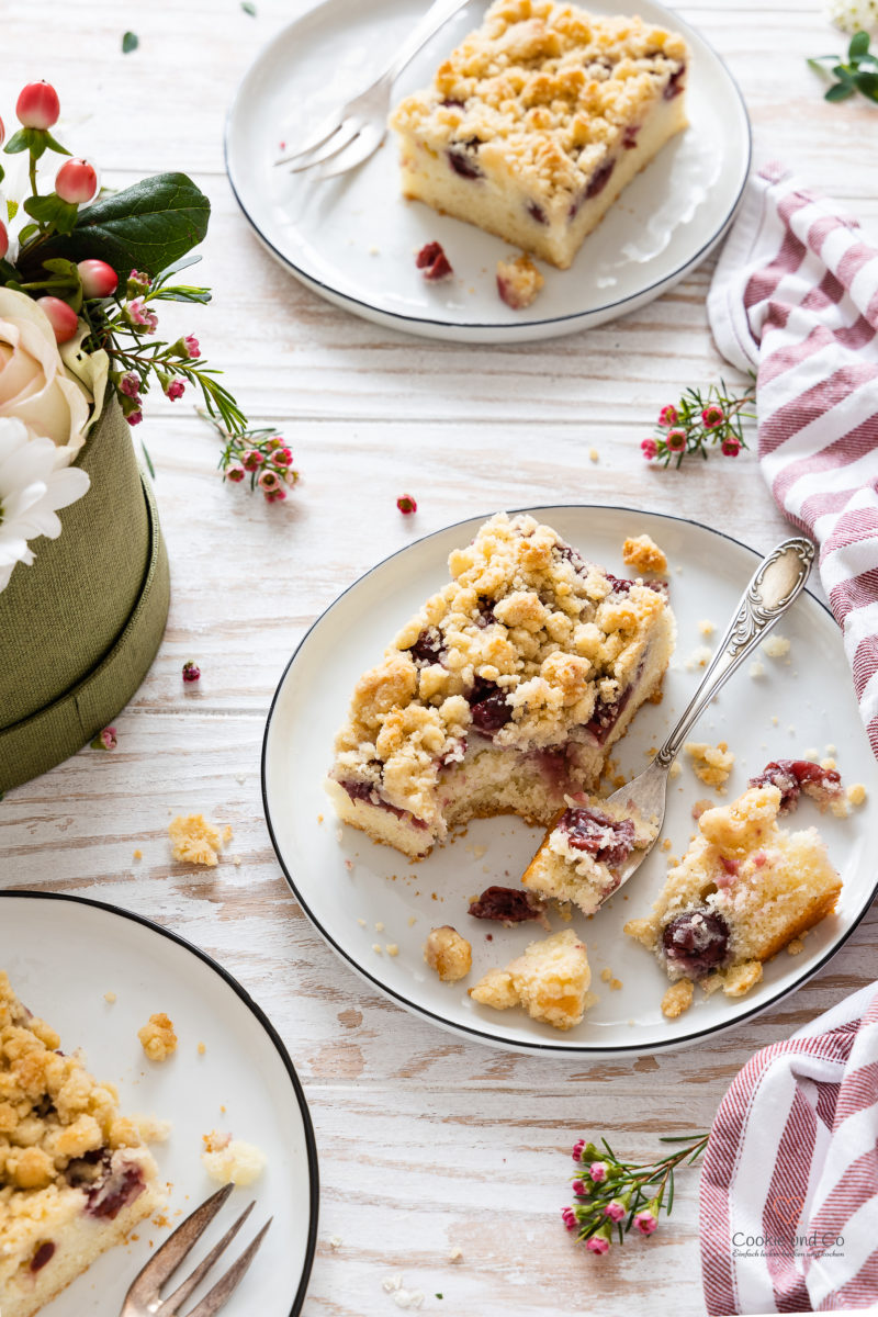 Ein Stück Kirsch-Streuselkuchen auf einem Teller mit einer Gabel und Blumen im Hintergrund (schneller Blechkuchen aus Rührteig)