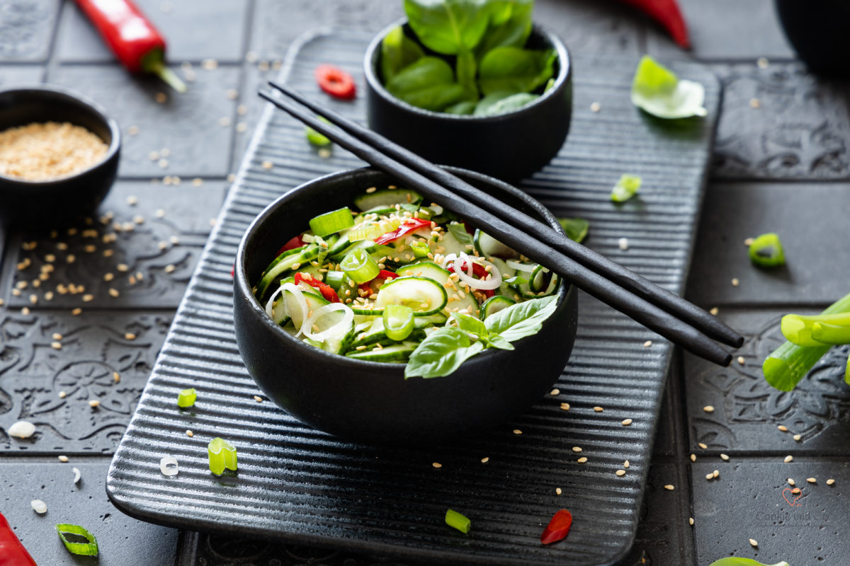 Thailändischer Gurkensalat (Ajad) in einer Schüssel mit Essstäbchen und Thai Basilikum im Hintergrund