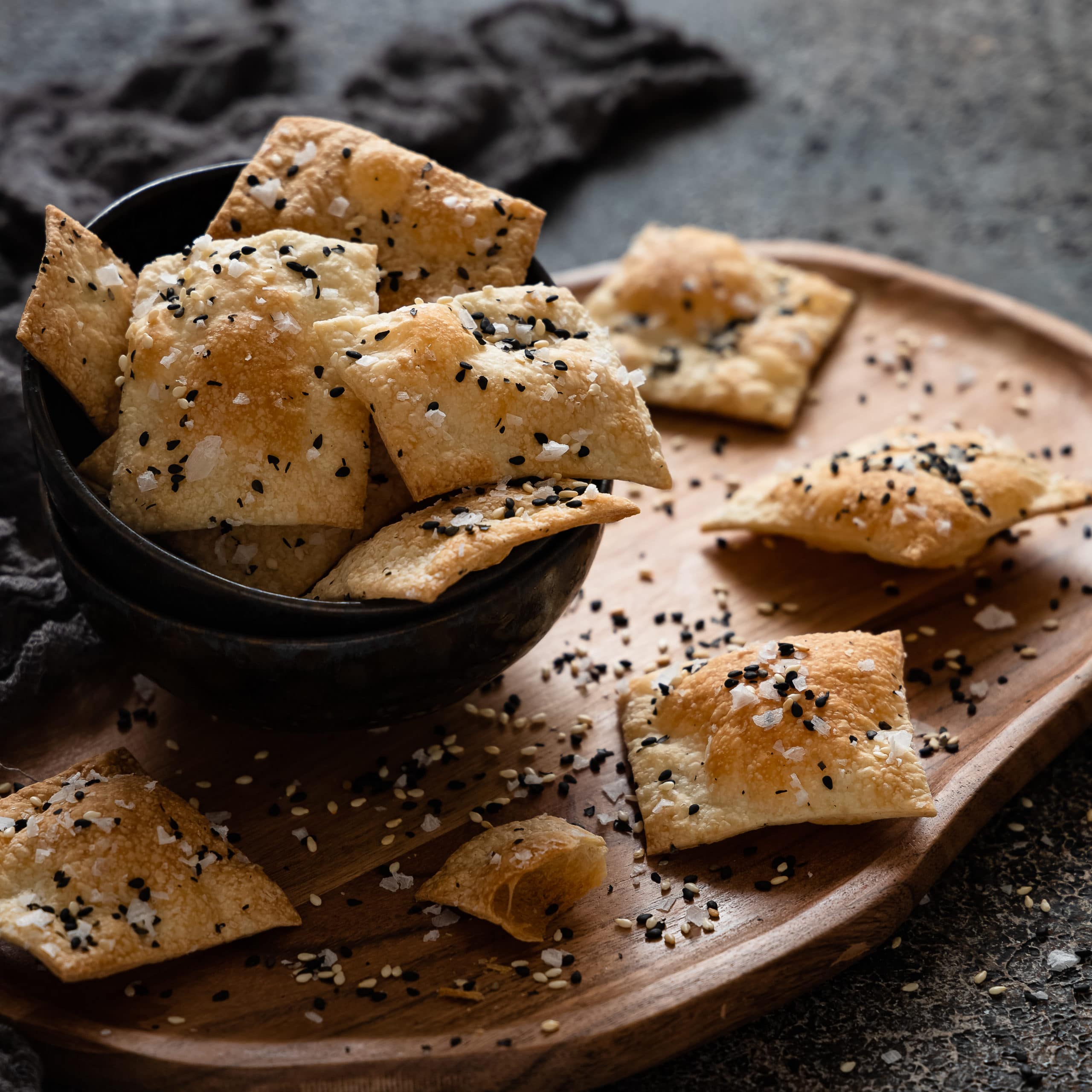 Knusprige Cracker mit Sesam in einer Schale auf einem Holbrett und einem Stofftuch im Hintergrund.