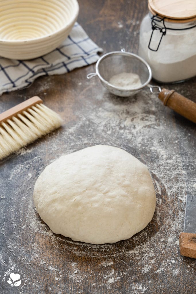 Ein vorgeformter Brotteig, während der Ballengare auf einem bemehlten Holztisch. mit Backutensilien drumherum.