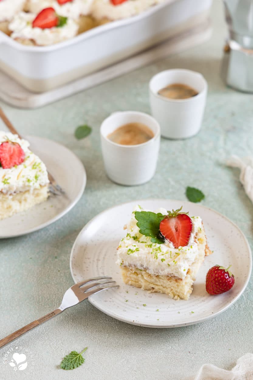 Ein Stück mexikanischer Tres leche Kuchen (Milchkuchen) mit Erdbeeren und Melisse auf einem Teller, einer Gabel im Vordergrund und einer Backform mit Kuchen und Espressotassen im Hintergrund.