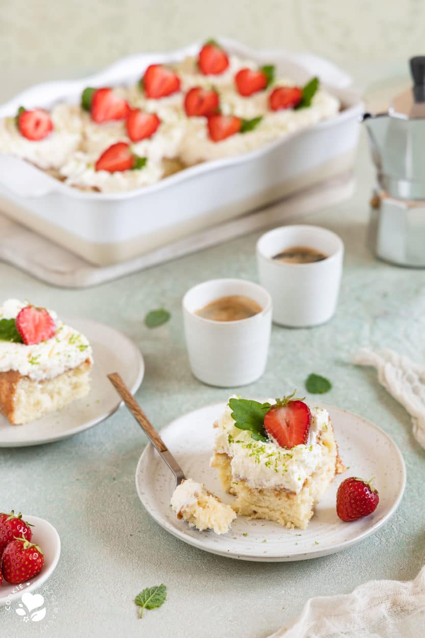Ein Stück mexikanischer Tres leche Kuchen (Milchkuchen) mit Erdbeeren auf einem Teller mit Espressotassen und einer Backform mit Kuchen im Hintergrund.