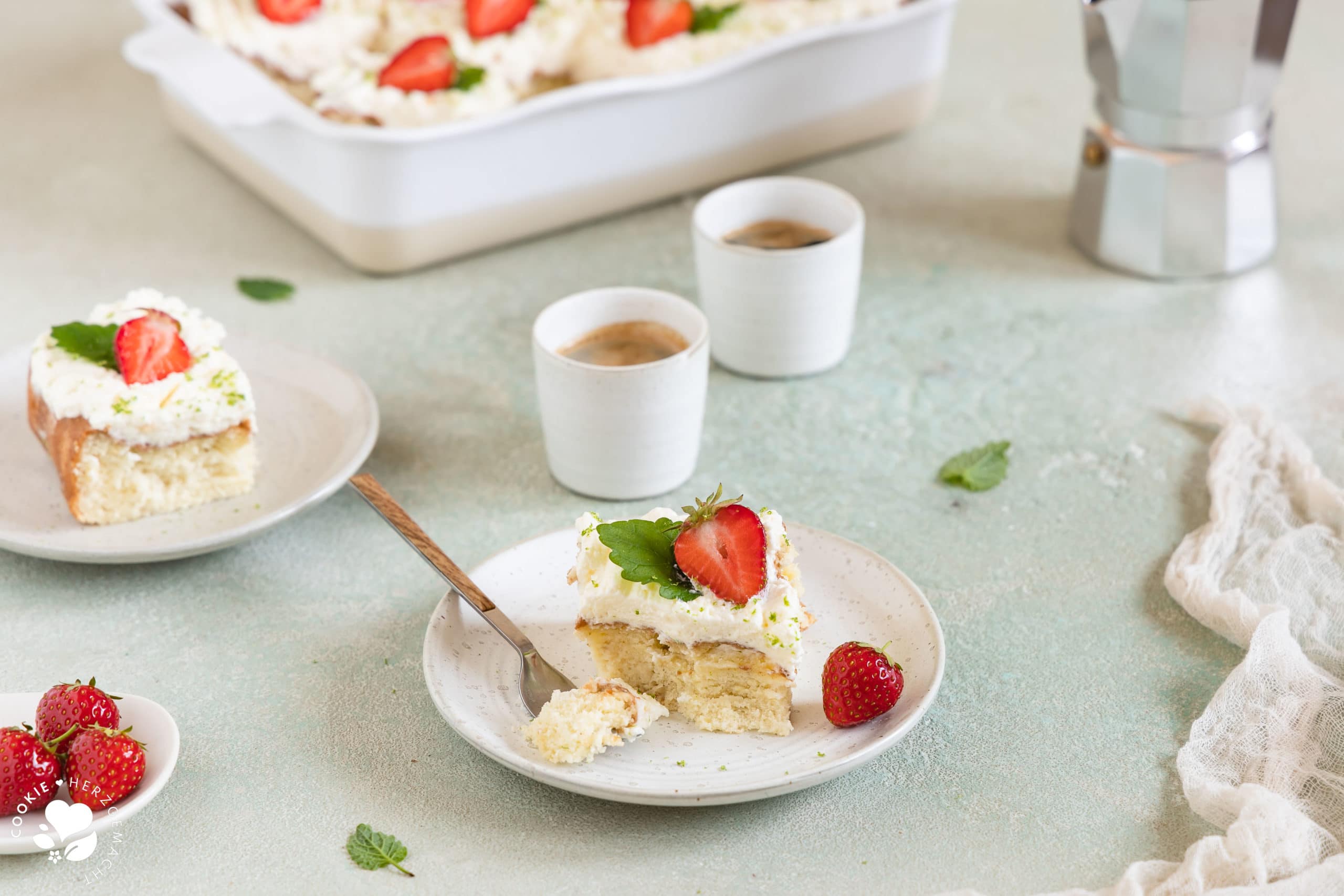 Ein Stück mexikanischer Tres leche Kuchen (Milchkuchen) mit Erdbeeren auf einem Teller mit Espressotassen und einer Backform mit Kuchen im Hintergrund.