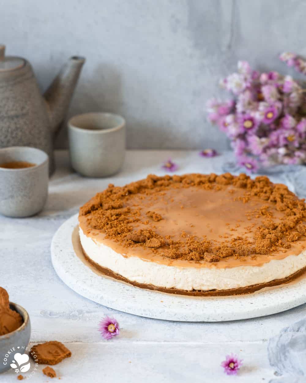 Karamell-Käsekuchen ohne Backen auf einer Kuchenplatte neben einer Schale Lotuskeksen und Kaffeetassen, einer Kaffeekanne und Blüten im Hintergrund.