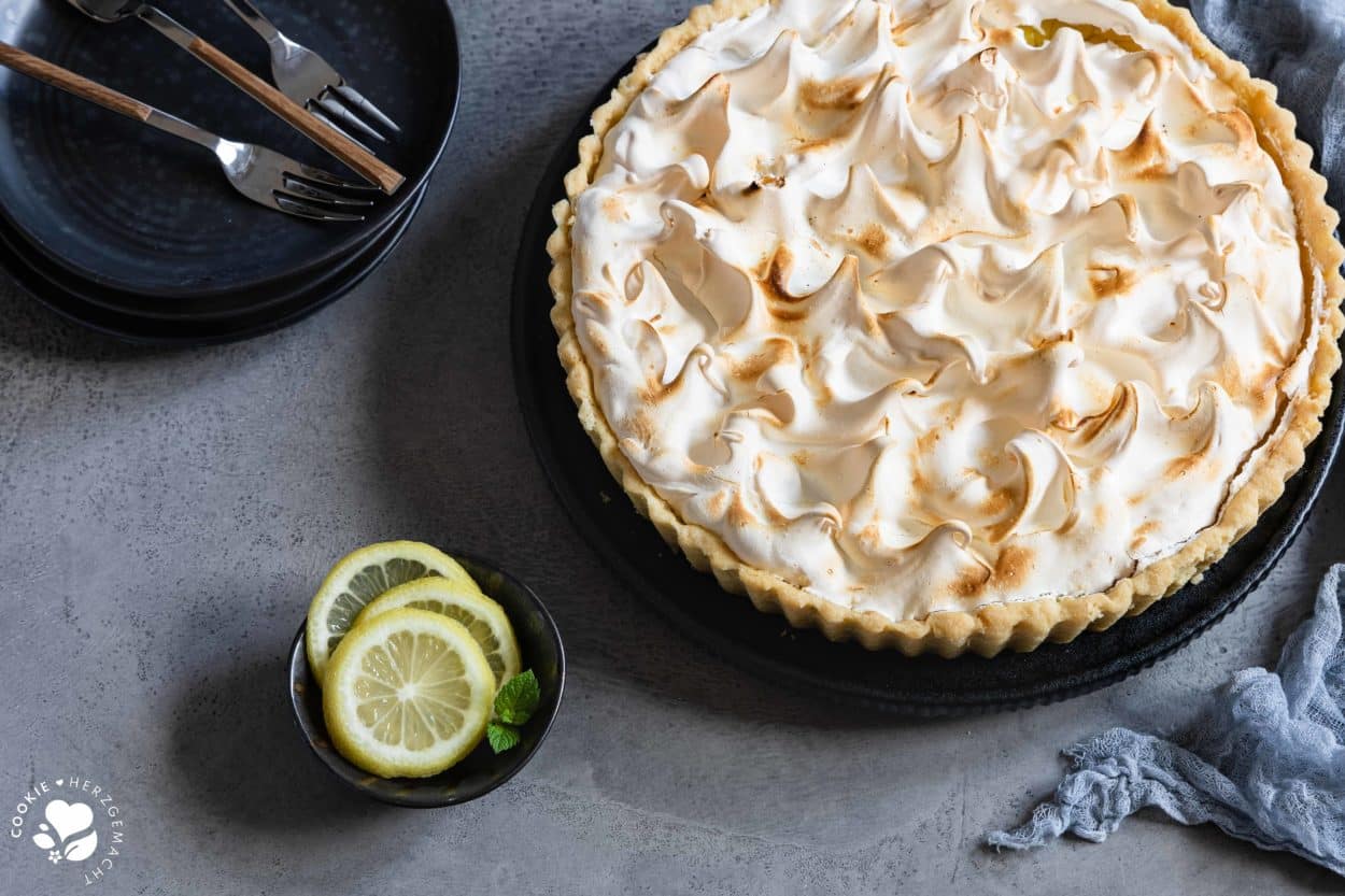 Zitronentarte mit Baiser auf einem Teller, einer Schale mit Zitronenscheiben und einem Stapel Tellern mit Kuchengabeln im Hintergrund.