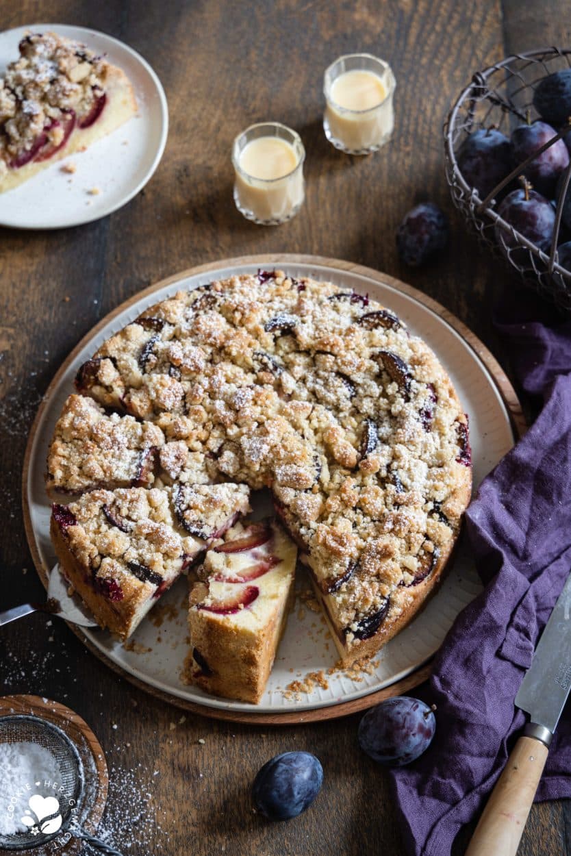Ein Zwetschgenkuchen mit Streuseln auf einer Tortenplatte mit zwei Gläsern Eierlikör im Hintergrund und einem Korb mit Pflaumen.