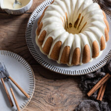 Ein Gugelhupf mit Zuckerguss auf einem Teller mit Zimtstangen und einem Kuchenteller mit Kuchengabeln im Vordergrund.