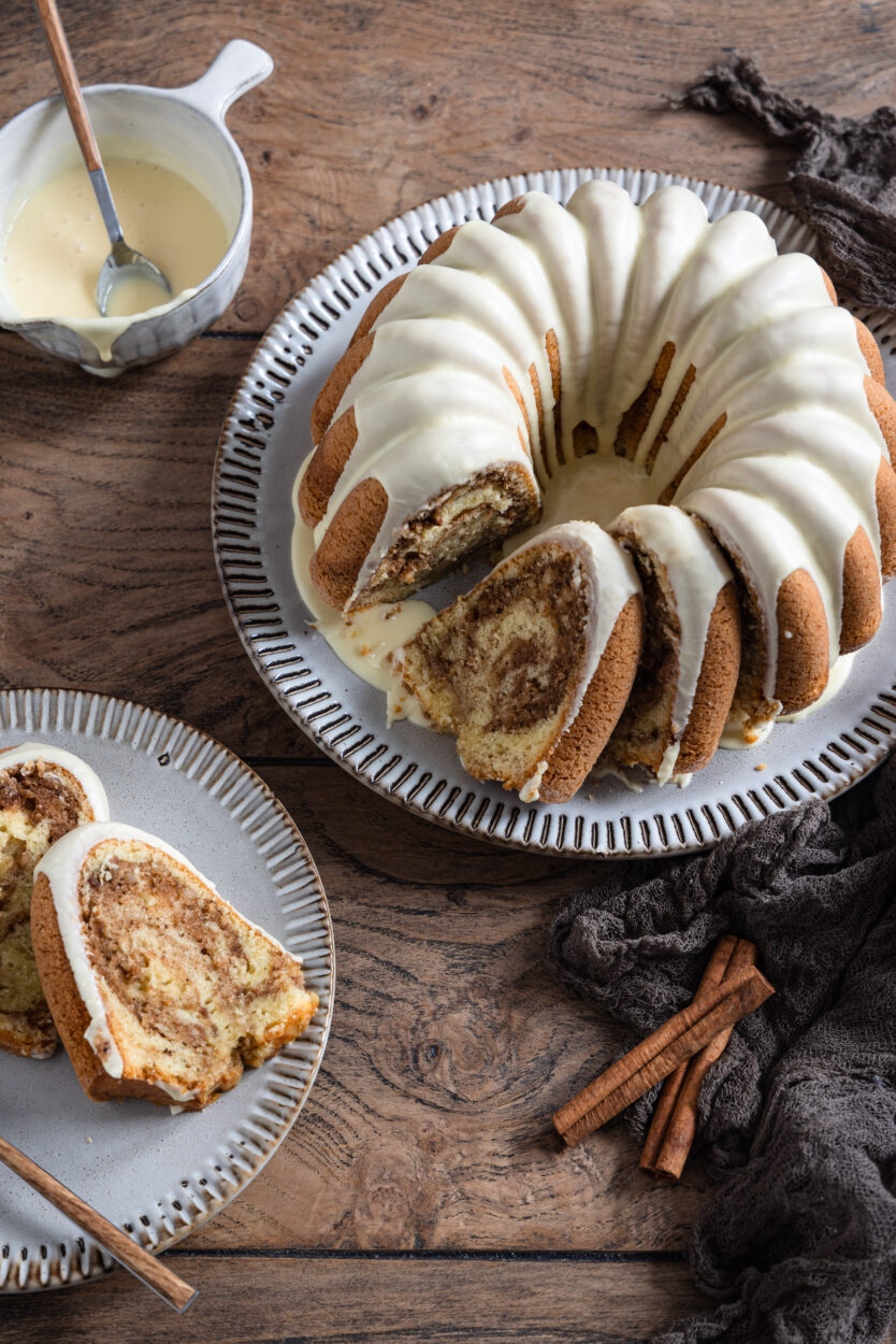 Ein angeschnittener Gugelhupf mit Zuckerguss auf einem Teller mit Zimtstangen und einem Kuchenteller mit Kuchenstücken im Vordergrund.