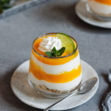 Schichtdessert mit Mango und Jog´hurt in einem Glas auf einem Teller mit einem Löffel.