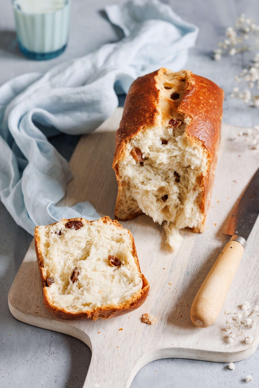 Rosinenstuten aufgeschnitten auf einem Holzbrett, mit einem Messer daneben und einem Glas Milch im Hintergrund.