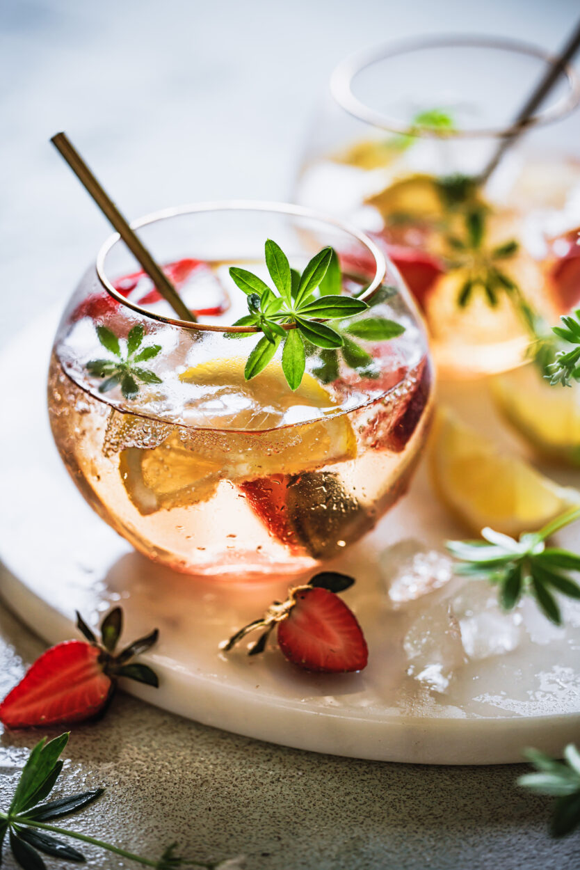 Maibowle mit Erdbeeren, Zitrone und Waldmeister in zwei Bowlegläsern auf einem Marmortablett mit Waldmeister und Erdbeeren im Vordergrund.