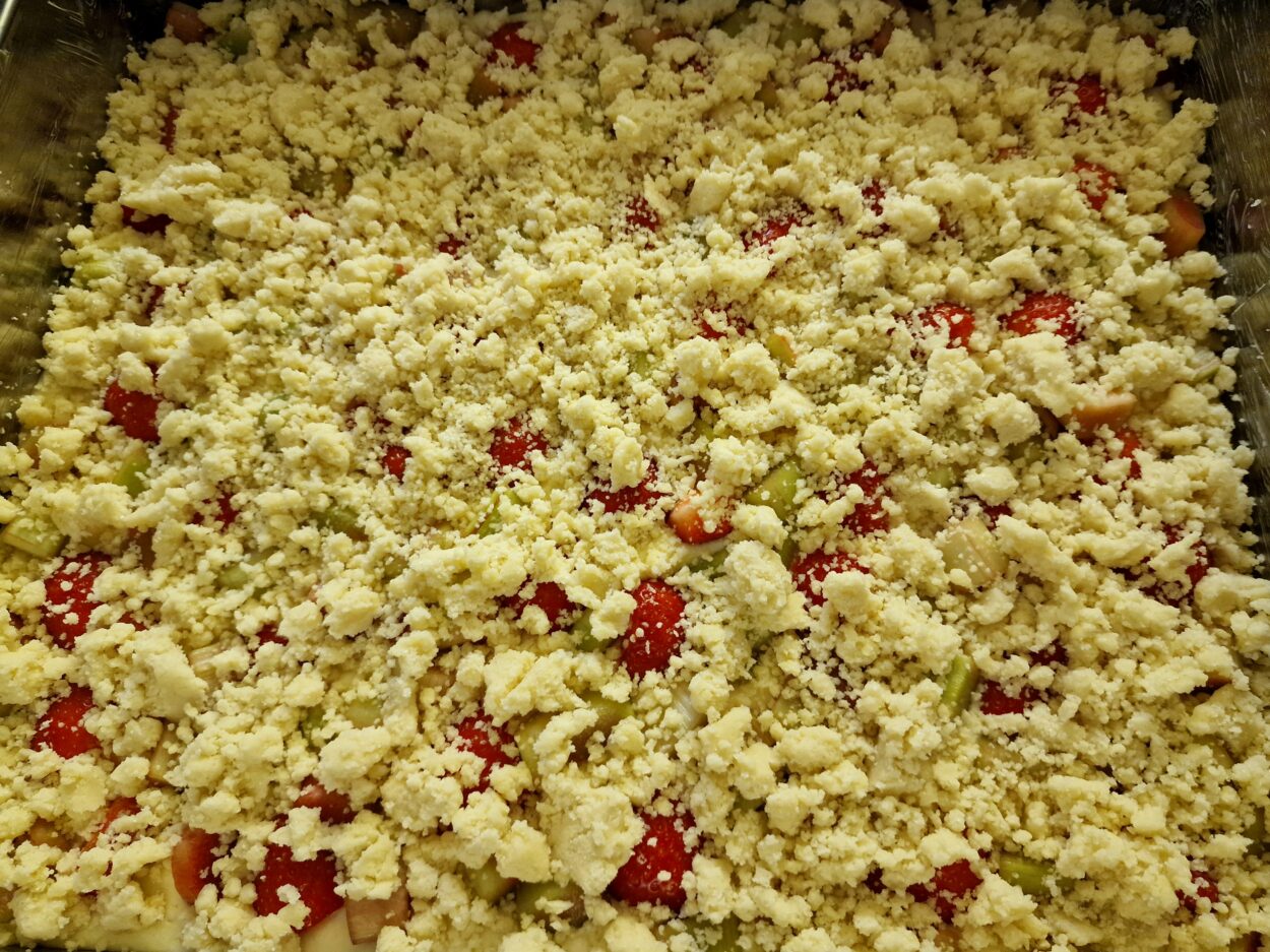 Streuselnteig über Erdbeeren und Rhabarberstücken auf Rührteig in einem Backrahmen vor dem Backen.