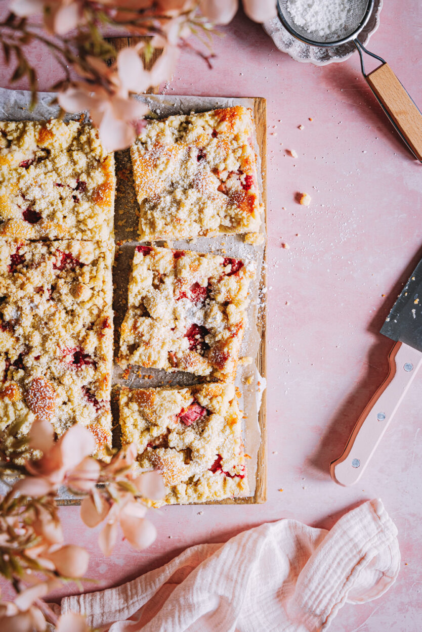 Erdbeer-Rhabarber-Kuchen mit Streuseln auf einem Brett mit einem Messer und einem Sieb mit Puderzucker daneben. 