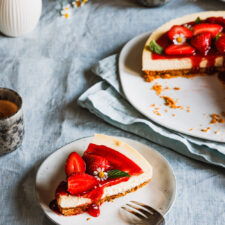Ein Stück Erdbeer-Cheesecake auf einem Teller mit zwei Tassen Kaffee daneben und dem ganzen Käsekuchen und Blüten im Hintergrund.
