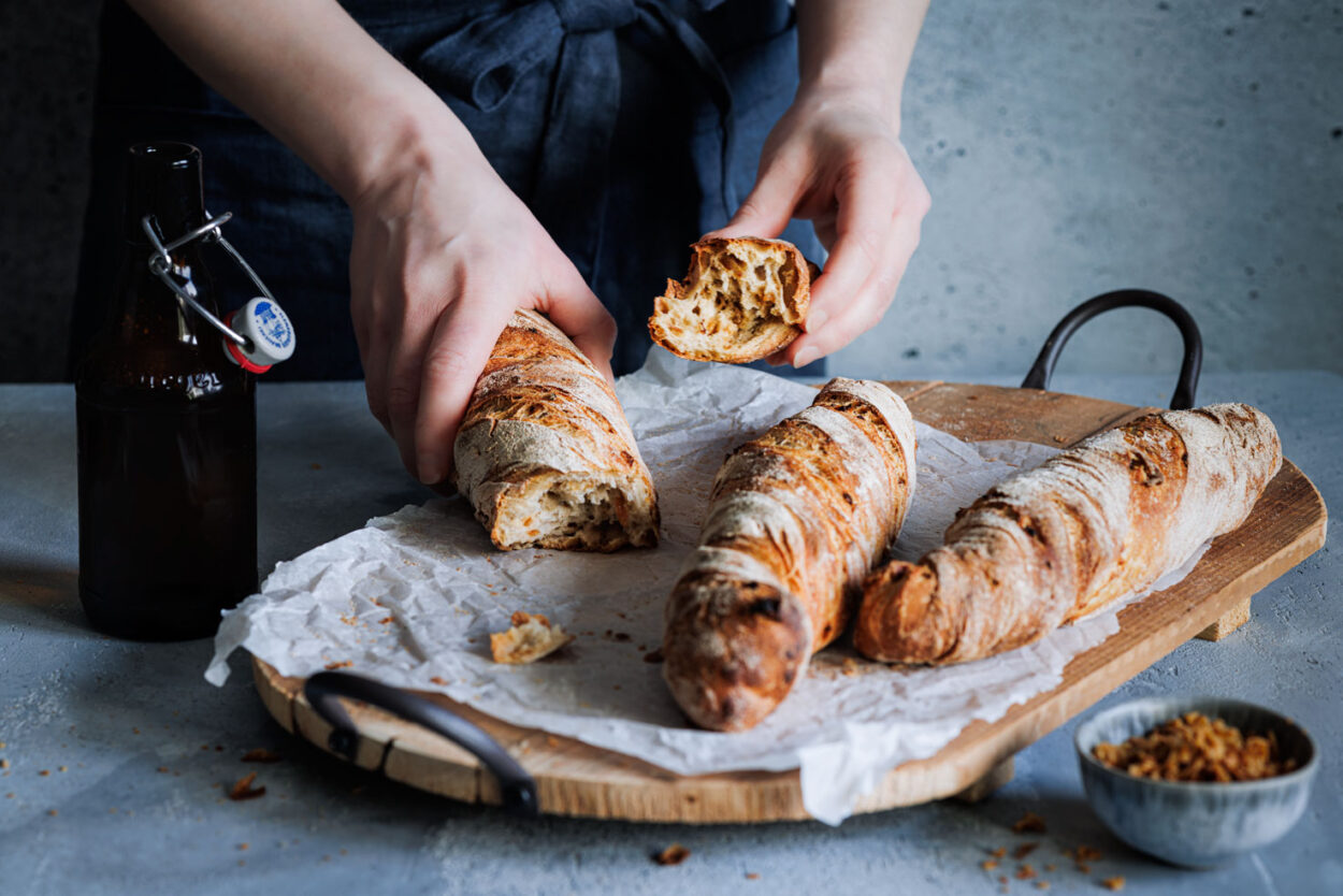 Zwiebel-Wurzelbrot auf einem rustikalen Holzbrett mit einer Flasche Bier im Hintergrund und einem Schälchen mit Röstzwiebeln im Vordergrund. 
