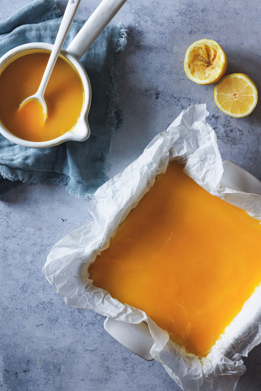 Zitronenkäsekuchen in einer Kuchenform mit Zitronen und einem Zopf mit dem Zitronentopping daneben. 