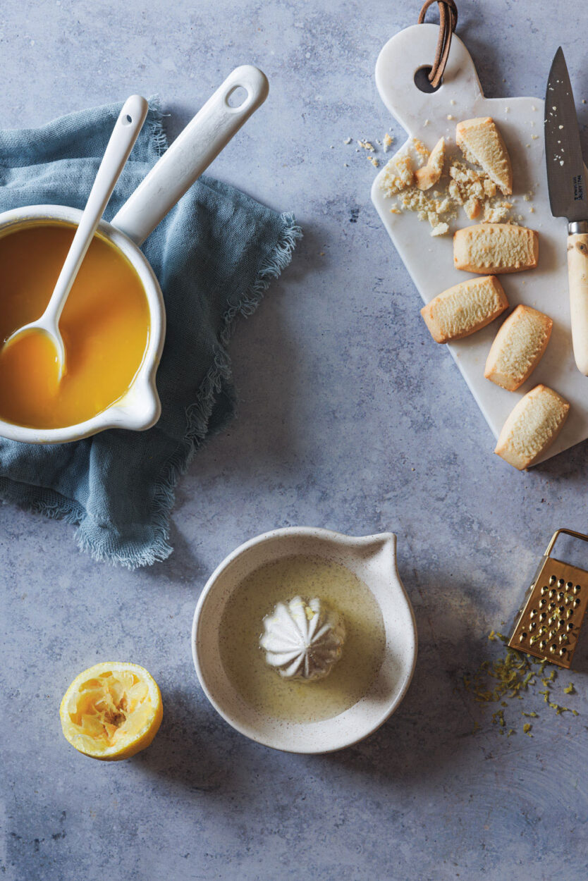 verschiedene Zutaten für einen Lemon Cheesecake wie Kekse, Zitronensaft, Zitronenschale und ein Topf einem Zitronenguss. 