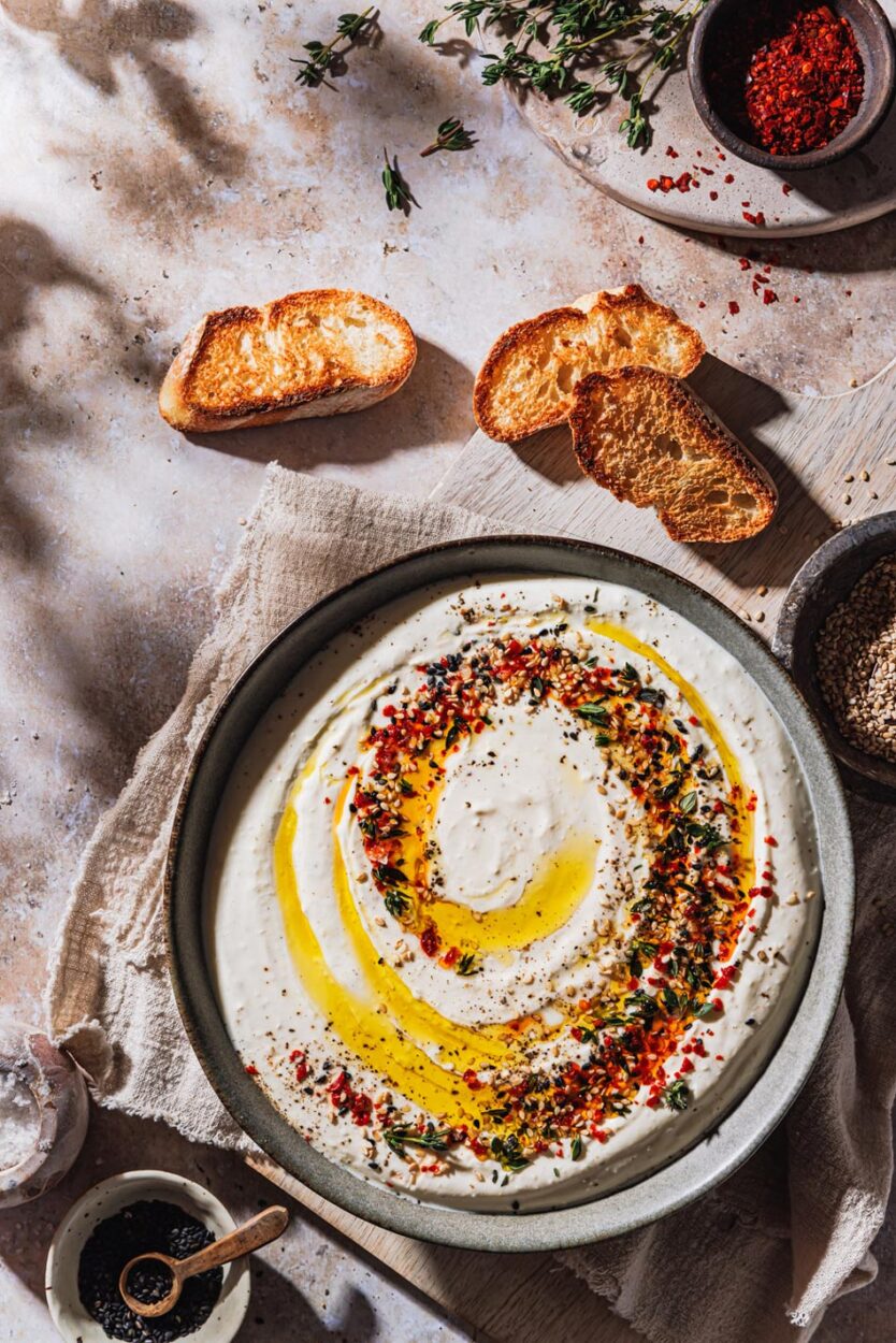 Feta-Dip in einer Schüssel mit Toppings sowie Schälchen mit Gewürzen und geröstetem Brot daneben.