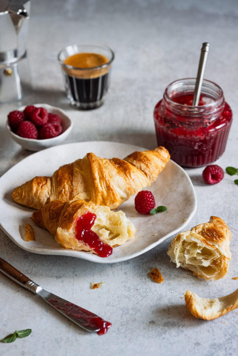 Himbeer-Marmelade ohne Kerne und Croissants auf einem Teller mit einem Marmeladenglas, einem Schälchen Himbeeren und Espresso im Hintergrund. 