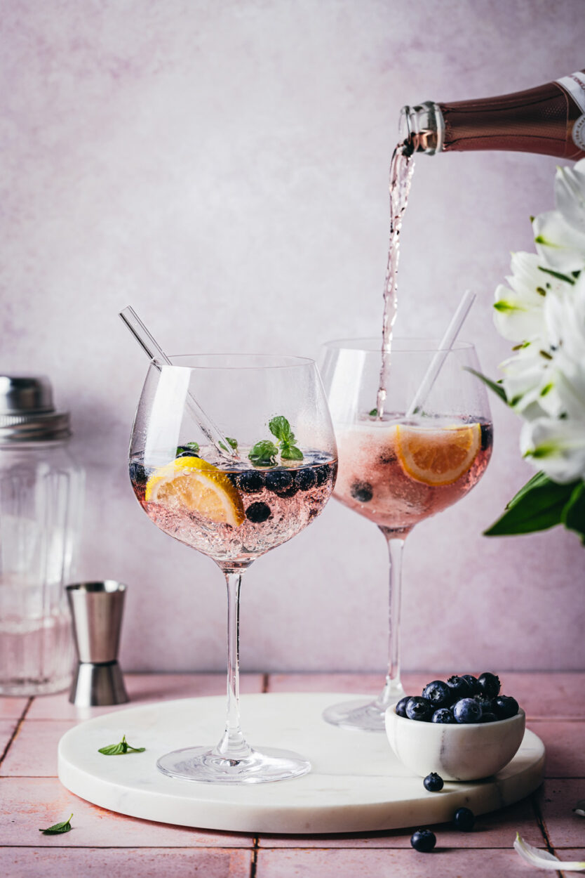 Heidelbeer-Cocktail in zwei Gläsern auf einer Marmorplatte mit weißen Blumen und frischen Heidelbeeren im Vordergrund. Im Hintergrund stehen ein Jigger und ein Cocktailshaker.