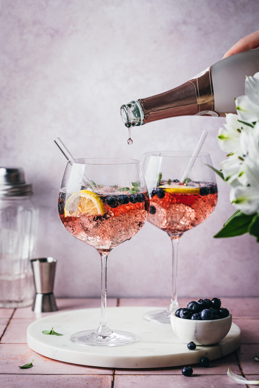 Blueberry Gin Spritz in zwei Cocktailgläsern auf einer Marmorplatte mit weißen Blumen und frischen Heidelbeeren im Vordergrund. Im Hintergrund stehen ein Jigger und ein Cocktailshaker.