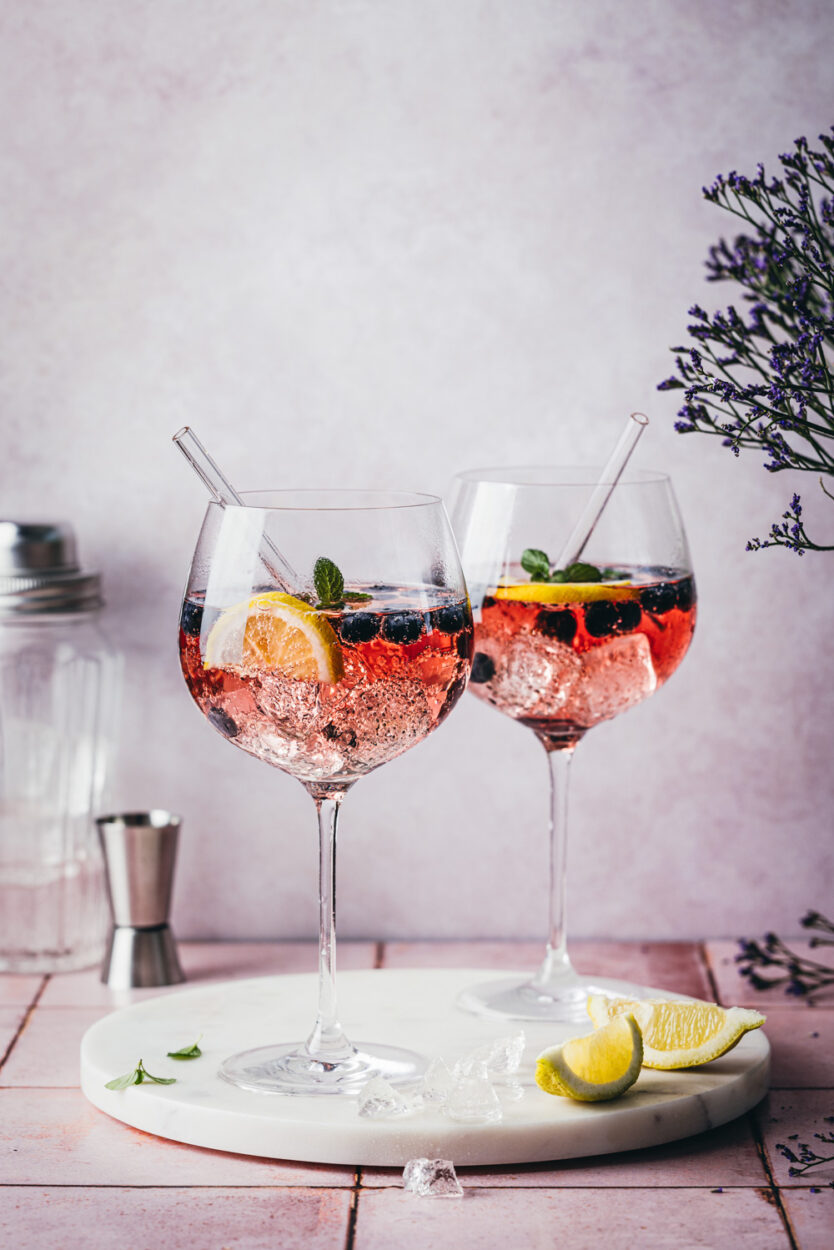 Blueberry Gin Spritz in zwei Cocktailgläsern auf einer Marmorplatte mit violetten Blumen und Zitronenspalten im Vordergrund. Im Hintergrund stehen ein Jigger und ein Cocktailshaker.