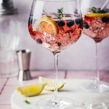 Blueberry Gin Spritz in zwei Cocktailgläsern auf einer Marmorplatte mit wZitronenspalten und Eiswürfeln im Vordergrund. Im Hintergrund stehen ein Jigger und ein Cocktailshaker.