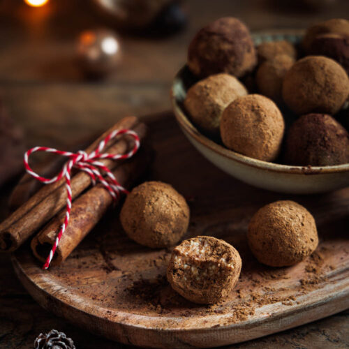 Spekulatiuskonfekt auf rustikalem Holzbrett angerichtet, dekoriert mit Zimtstangen und weihnachtlicher Beleuchtung im Hintergrund – perfekt für die Festtage.